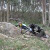 Vestidos con equipo de paintball, dos participantes se agachan detrás de grandes rocas en una zona boscosa, disfrutando plenamente de la emoción de "La mejor experiencia de paintball en A Coruña. - ForTwo - Planes para parejas