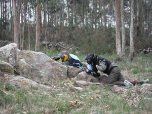 Vestidos con equipo de paintball, dos participantes se agachan detrás de grandes rocas en una zona boscosa, disfrutando plenamente de la emoción de "La mejor experiencia de paintball en A Coruña. - ForTwo - Planes para parejas