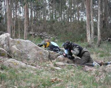 Vestidos con equipo de paintball, dos participantes se agachan detrás de grandes rocas en una zona boscosa, disfrutando plenamente de la emoción de "La mejor experiencia de paintball en A Coruña. - ForTwo - Planes para parejas