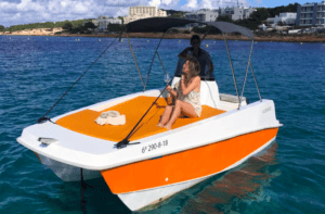 Dos personas se relajan en un barco naranja y blanco en las aguas cristalinas de una orilla de arena, abrazando el espíritu de "Navega Ibiza a tu Ritmo: Barcos sin Licencia" mientras exploran sin límites. - ForTwo - Planes para parejas