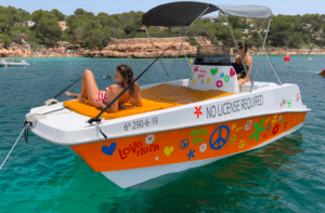 Una pequeña embarcación decorada con el lema "Navega Ibiza a tu Ritmo: Barcos sin Licencia" flota con dos personas a bordo en las cristalinas aguas de Ibiza. Disfruta de la libertad de explorar este impresionante paraíso sin necesidad de licencia. - ForTwo - Planes para parejas