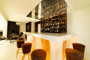 Este elegante bar en PAIISE Hotels Ibiza: Relájate cerca de las playas ofrece un elegante mostrador blanco y un techo con espejos, con estantes llenos de botellas cálidamente rodeados de taburetes marrones, que invitan tanto a los aventureros de la isla como a los amantes de la playa. - ForTwo - Planes para parejas