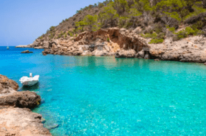 En "Paddle surf en el norte de Ibiza", las cristalinas aguas turquesas de una cala rocosa se enmarcan entre frondosos árboles. Un pequeño barco está amarrado a la izquierda, mientras que las suaves olas invitan a la aventura del paddle surf en este tranquilo paraíso. - ForTwo - Planes para parejas