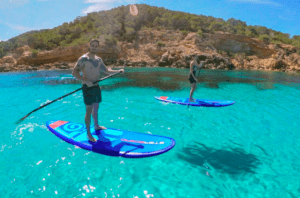 Dos personas disfrutan del Paddle surf en el norte de Ibiza en claras aguas turquesas cerca de la orilla rocosa y arbolada bajo un cielo soleado. - ForTwo - Planes para parejas