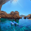 Dos personas se encuentran sobre sus tablas de Paddle surf en el norte de Ibiza en las cristalinas aguas azules, tomadas de la mano debajo de un arco rocoso bajo un cielo azul brillante. - ForTwo - Planes para parejas