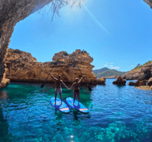 Dos personas se encuentran sobre sus tablas de Paddle surf en el norte de Ibiza en las cristalinas aguas azules, tomadas de la mano debajo de un arco rocoso bajo un cielo azul brillante. - ForTwo - Planes para parejas