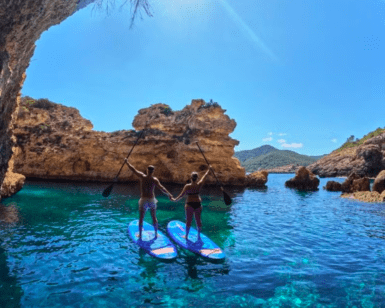 Dos personas se encuentran sobre sus tablas de Paddle surf en el norte de Ibiza en las cristalinas aguas azules, tomadas de la mano debajo de un arco rocoso bajo un cielo azul brillante. - ForTwo - Planes para parejas