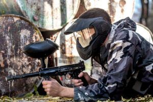 Paintball "El Superviviente" en Pontevedra muestra a un jugador camuflado con una máscara apuntando hábilmente con una pistola de paintball, agachado junto a barriles de madera al aire libre. - ForTwo - Planes para parejas