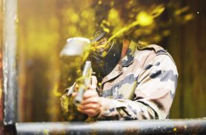 En los emocionantes campos de batalla de Pontevedra, un jugador se pone equipo de camuflaje y máscara para una intensa ronda de Paintball "El Superviviente", disparando una pistola de paintball mientras pintura amarilla vibrante salpica alrededor. - ForTwo - Planes para parejas