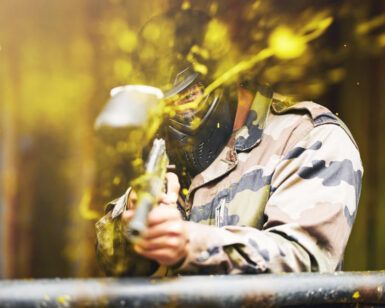 En los emocionantes campos de batalla de Pontevedra, un jugador se pone equipo de camuflaje y máscara para una intensa ronda de Paintball "El Superviviente", disparando una pistola de paintball mientras pintura amarilla vibrante salpica alrededor. - ForTwo - Planes para parejas