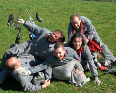 Un grupo de hombres con uniformes iguales, riendo y jugando, amontonados unos sobre otros en el césped de Asturias, con sus marcadores de Paintball en Asturias: ¡Juegos de guerra en el bosque! esparcidos cerca, listos para más acción. - ForTwo - Planes para parejas