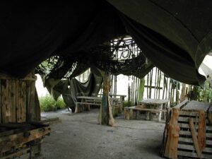 La experiencia Paintball en Asturias ofrece un refugio rústico con bancos y mesas de madera bajo un dosel de tela verde, ideal para trazar estrategias antes de una emocionante partida de paintball en un exuberante entorno forestal. - ForTwo - Planes para parejas