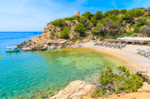Una playa aislada de aguas turquesas, arena y tumbonas se encuentra entre acantilados rocosos y vegetación. Perfecta para un paseo en barco a Es Vedrá y disfrutar de las vistas de Es Vedrá elevándose majestuosamente sobre el mar resplandeciente. - ForTwo - Planes para parejas