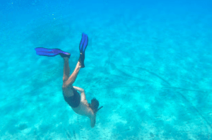 Una persona que practica snorkel en las cristalinas aguas turquesas con aletas azules se deleita con la impresionante vista de Es Vedrá, como si estuviera experimentando un mágico "Paseo en barco a Es Vedrá" bajo el mar. - ForTwo - Planes para parejas