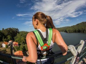 En el puente de Pontevedra, una mujer con un arnés de seguridad de "Puenting en Pontevedra, en Sinde" contempla el río Sinde y el frondoso bosque bajo un cielo despejado. - ForTwo - Planes para parejas