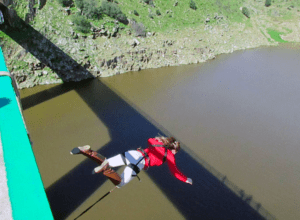 Ataviado con una chaqueta roja y un arnés, la persona salta valientemente desde un puente sobre el río Guadiana, experimentando la emocionante emoción de ¡Atrévete a Volar sobre el Guadiana! mientras parece volar por el aire libre. - ForTwo - Planes para parejas