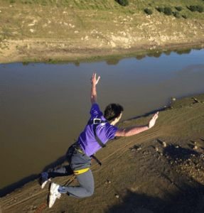 Un atrevido entusiasta se lanza con los brazos extendidos sobre el río Guadiana, luciendo una vibrante camisa y arnés de color morado en la experiencia Puenting ¡Atrévete a Volar sobre el Guadiana!. - ForTwo - Planes para parejas