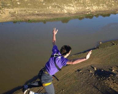 Un atrevido entusiasta se lanza con los brazos extendidos sobre el río Guadiana, luciendo una vibrante camisa y arnés de color morado en la experiencia Puenting ¡Atrévete a Volar sobre el Guadiana!. - ForTwo - Planes para parejas