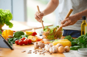 Una persona está inmersa en un Taller de Cocina Saludable, preparando hábilmente una ensalada con verduras frescas sobre una encimera de madera, mientras cerca una tableta muestra recetas para inspirarse. - ForTwo - Planes para parejas