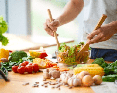 Una persona está inmersa en un Taller de Cocina Saludable, preparando hábilmente una ensalada con verduras frescas sobre una encimera de madera, mientras cerca una tableta muestra recetas para inspirarse. - ForTwo - Planes para parejas