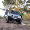 Dos personas posan juguetonamente sobre un todoterreno elevado estacionado en un terreno rocoso, rodeado de árboles, durante su aventura Tour en 4x4 desde La Coruña. - ForTwo - Planes para parejas