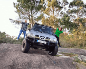 Dos personas posan juguetonamente sobre un todoterreno elevado estacionado en un terreno rocoso, rodeado de árboles, durante su aventura Tour en 4x4 desde La Coruña. - ForTwo - Planes para parejas