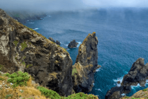 Acantilados costeros escarpados se encuentran con un mar brumoso bajo un cielo nublado cerca de La Coruña, coronado por una exuberante vegetación, ideal para el inolvidable Tour en 4x4 desde La Coruña. - ForTwo - Planes para parejas