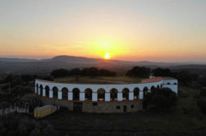 El "Tour en 4x4 por los Llanos de Olivenza" te lleva a un edificio ovalado con ventanas y azotea arqueadas, rodeado de árboles al atardecer con vistas a la montaña, un lugar ideal para explorar una belleza serena y paisajes impresionantes. - ForTwo - Planes para parejas