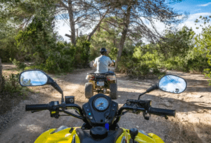 Un motociclista en un quad amarillo sigue a otro a través de un sendero forestal, rodeado de árboles y el cielo azul: una experiencia emocionante similar al "Tour en quad por Ibiza: Aventura y paisajes únicos". - ForTwo - Planes para parejas