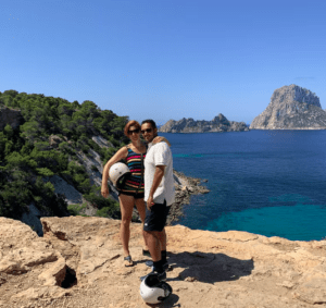 Una pareja con cascos posa sobre un acantilado rocoso, con vistas al mar y capturando la esencia del "Tour en quad por Ibiza: Aventura y paisajes únicos", con una gran formación rocosa al fondo. - ForTwo - Planes para parejas