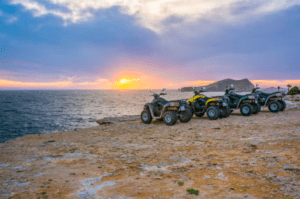 Experimenta la emoción de un "Tour en quad por Ibiza: Aventura y paisajes únicos" con cuatro ATV en un acantilado rocoso al atardecer, con vistas al mar y a una isla lejana bajo un cielo colorido, capturando el vibrante encanto de Ibiza. - ForTwo - Planes para parejas