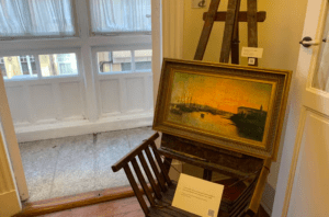 Un cuadro enmarcado reposa sobre un caballete de madera junto a una ventana iluminada por el sol, junto a una silla y una placa, capturando la esencia de Visita la Casa-Museo de Picasso. - ForTwo - Planes para parejas