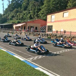 Personas en karts en el autódromo de Soto de Dueñas, preparadas para correr, con un edificio y árboles al fondo, encarnan la emoción del karting en "Vive la velocidad en el Karting Soto de Dueñas, Asturias. - ForTwo - Planes para parejas