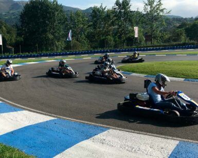 En Karting Soto de Dueñas, Asturias, los competidores compiten con maestría en karts en una pista con bordes azules y blancos, rodeada de frondosos árboles y montañas impresionantes. Experimenta la emoción de "Vive la velocidad". - ForTwo - Planes para parejas