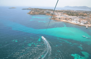 Desde arriba, una embarcación se desliza por las aguas turquesas de Santa Eulalia del Río, flanqueada por edificios costeros y exuberante vegetación. Cerca de allí, los aventureros disfrutan de la emoción del parasailing con Vuelo en Parasailing en Santa Eulalia del Río. - ForTwo - Planes para parejas