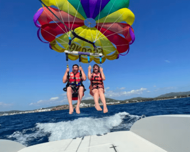 Dos personas disfrutan de una emocionante experiencia de Vuelo en Parasailing en Santa Eulalia del Río, volando alto sobre el océano con un paracaídas vibrante y usando chalecos salvavidas. - ForTwo - Planes para parejas