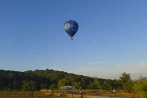 Vuelo en globo: Ibiza desde las alturas al amanecer" ofrece un vuelo sereno en un globo aerostático azul, que se desplaza con gracia sobre el paisaje rural de Ibiza, donde árboles y colinas se despliegan bajo el cielo despejado. - ForTwo - Planes para parejas