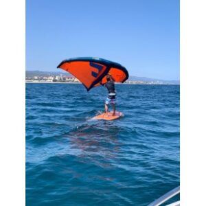 Bajo un cielo despejado, una persona practica wingfoil sobre aguas azules con las lejanas Costas como impresionante telón de fondo. "Wingfoil: La Nueva Sensación de las Costas" encarna a la perfección la aventura y la libertad en cada ola. - ForTwo - Planes para parejas