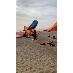 Una persona en la playa sostiene alegremente una cometa naranja y azul, rodeada de cometas dispersas y equipo Wingfoil: La Nueva Sensación de las Costas. Esta vibrante escena captura la mezcla de aventura y serenidad bajo el sol. - ForTwo - Planes para parejas