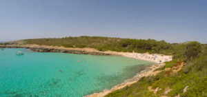 Una playa tranquila en Mallorca, con aguas turquesas y exuberante vegetación, acoge a algunos bañistas y toma el sol. Cerca, un "Barco sin Licencia en Mallorca" flota tranquilamente sobre las suaves olas, lo que realza la tranquilidad. - ForTwo - Planes para parejas