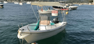 Una pequeña lancha con toldo está fondeada en el puerto de Mallorca, con un cartel de "Se alquila". Es perfecta para quienes buscan "Barco sin licencia en Mallorca", con varias otras embarcaciones visibles al fondo. Ideal para los amantes del alquiler de barcos. - ForTwo - Planes para parejas