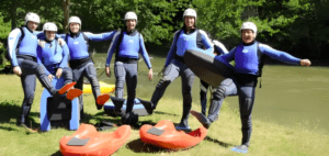 Seis personas con trajes de neopreno y casco posan con kayaks en la ribera herbosa del Alto Ebro, levantando una pierna y sonriendo con alegría. Su espíritu contagioso captura la aventura de Hidrospeed en el Alto Ebro, una experiencia emocionante. - ForTwo - Planes para parejas