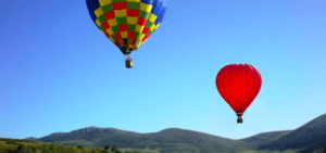 El Vuelo en Globo en Reinosa ofrece una experiencia fascinante con dos globos aerostáticos, uno multicolor y otro rojo, flotando sobre colinas verdes bajo un cielo azul claro. - ForTwo - Planes para parejas
