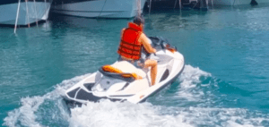 Una persona con un chaleco salvavidas rojo navega en una Alquila una Moto de Agua en Menorca por las cristalinas aguas azules, con barcos visibles al fondo, ideal para tu aventura de alquiler en un paisaje impresionante. - ForTwo - Planes para parejas