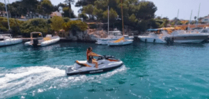 Una persona disfruta de un emocionante paseo en una "Alquila una Moto de Agua en Menorca" entre varios barcos y una exuberante vegetación en un pintoresco puerto de Menorca. - ForTwo - Planes para parejas