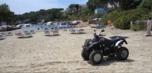 Un ATV en una playa de arena de Menorca, rodeado de tumbonas vacías y sombrillas junto al mar, crea una escena inolvidable para una "Aventura en Quad por Menorca. - ForTwo - Planes para parejas