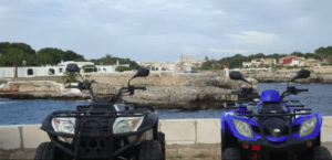 Dos quads aparcados frente al mar ofrecen una vista impresionante de la costa rocosa de Menorca y su encantadora ciudad, ideal para una Aventura en Quad por Menorca. - ForTwo - Planes para parejas