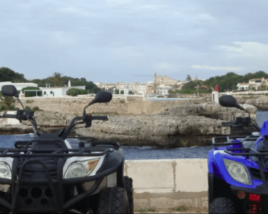 Dos quads aparcados frente al mar ofrecen una vista impresionante de la costa rocosa de Menorca y su encantadora ciudad, ideal para una Aventura en Quad por Menorca. - ForTwo - Planes para parejas