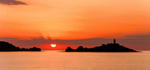 Una puesta de sol pinta el cielo de naranja sobre el tranquilo mar de Mallorca, con una isla distante y un faro recortados, evocando pensamientos de Avistamiento de Delfines en Mallorca en las serenas aguas de abajo. - ForTwo - Planes para parejas
