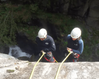 Dos amantes de las emociones fuertes, ataviados con cascos, descienden en rápel por un acantilado junto a una cascada, iniciando una emocionante aventura de Barranquismo de Iniciación en el Valle de Núria. - ForTwo - Planes para parejas